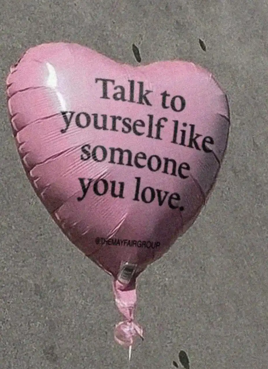 A pink heart-shaped balloon lies on gray pavement, printed with the text 'Talk to yourself like someone you love.' and a…