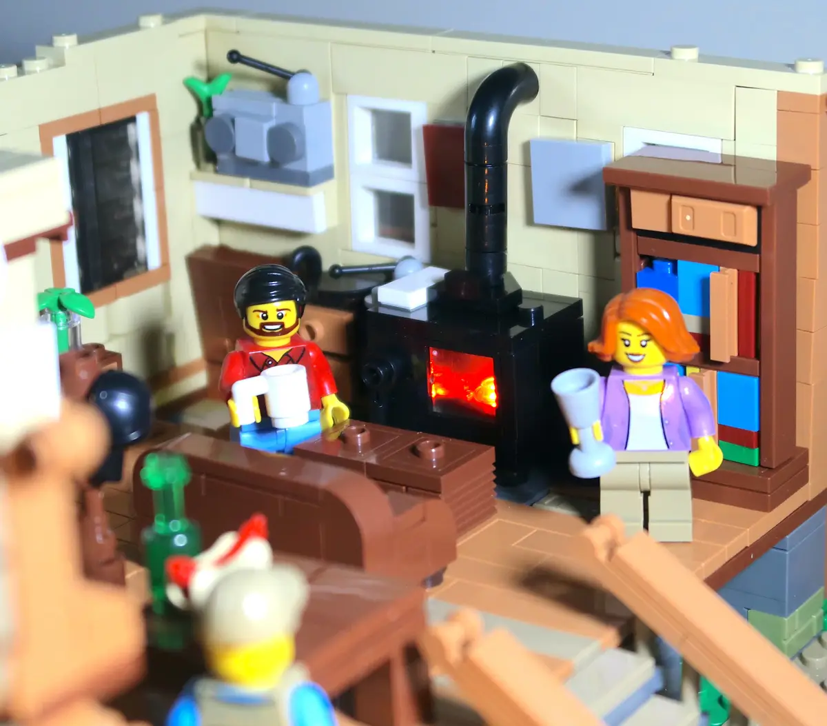 A Lego diorama depicts a cozy living room with two smiling Lego minifigures, a man in a red shirt and a woman in a purple…