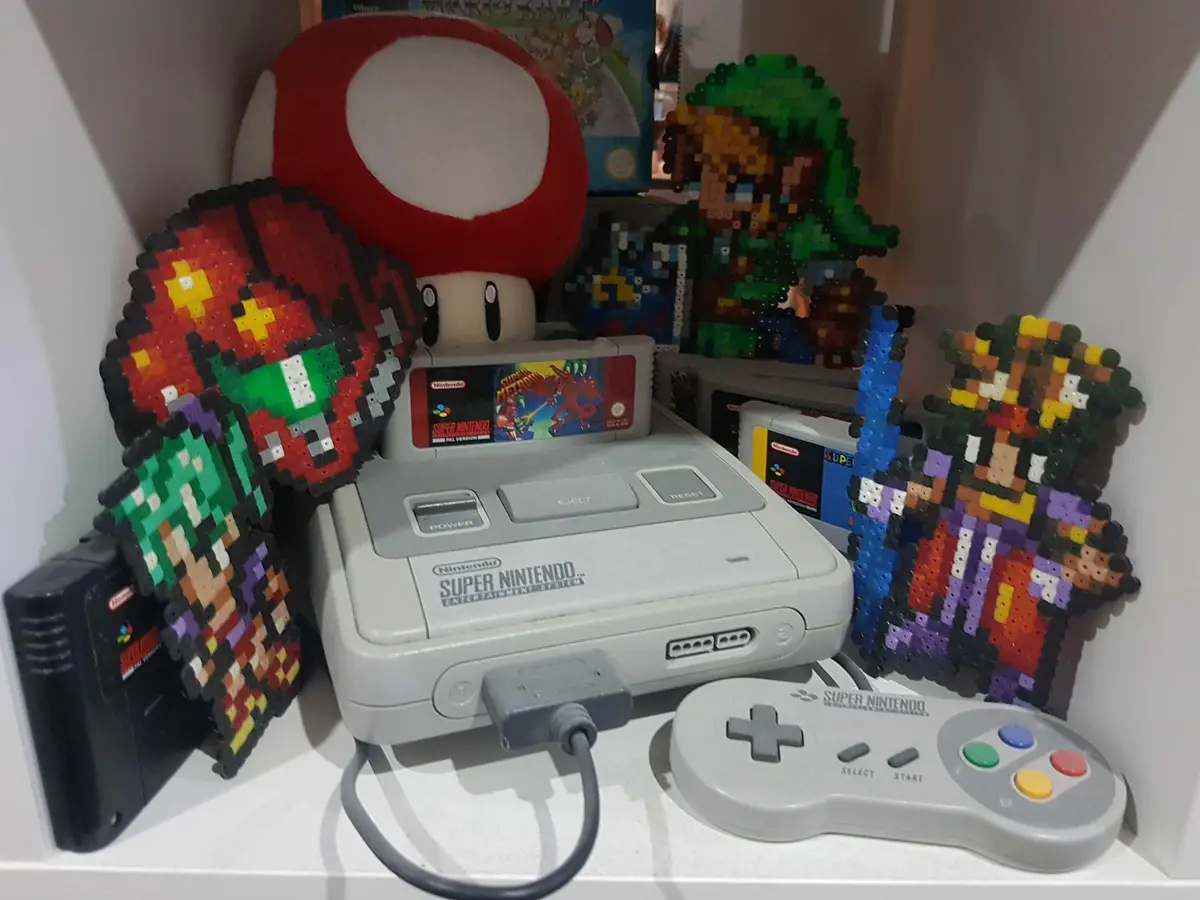A Super Nintendo Entertainment System console sits on a shelf surrounded by pixel-art perler bead figures of Mario, Luigi…