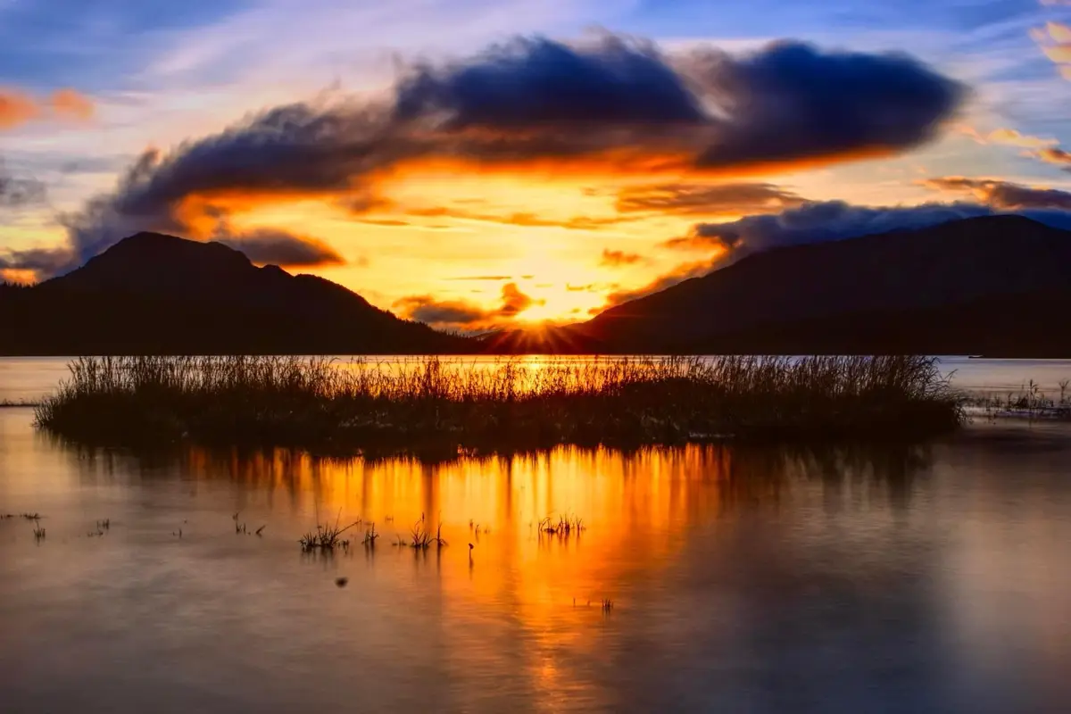 A serene landscape photo of a sunset over a lake with mountains in the background, featuring vibrant orange and yellow hues…