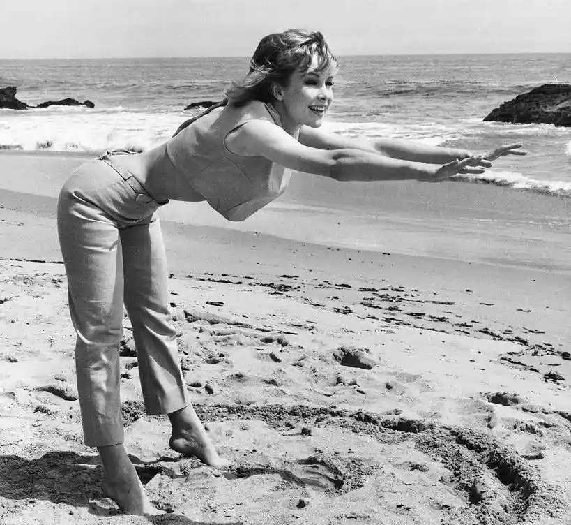 A black-and-white photo of a smiling woman on a beach, leaning forward with arms outstretched as if flying, while standing…