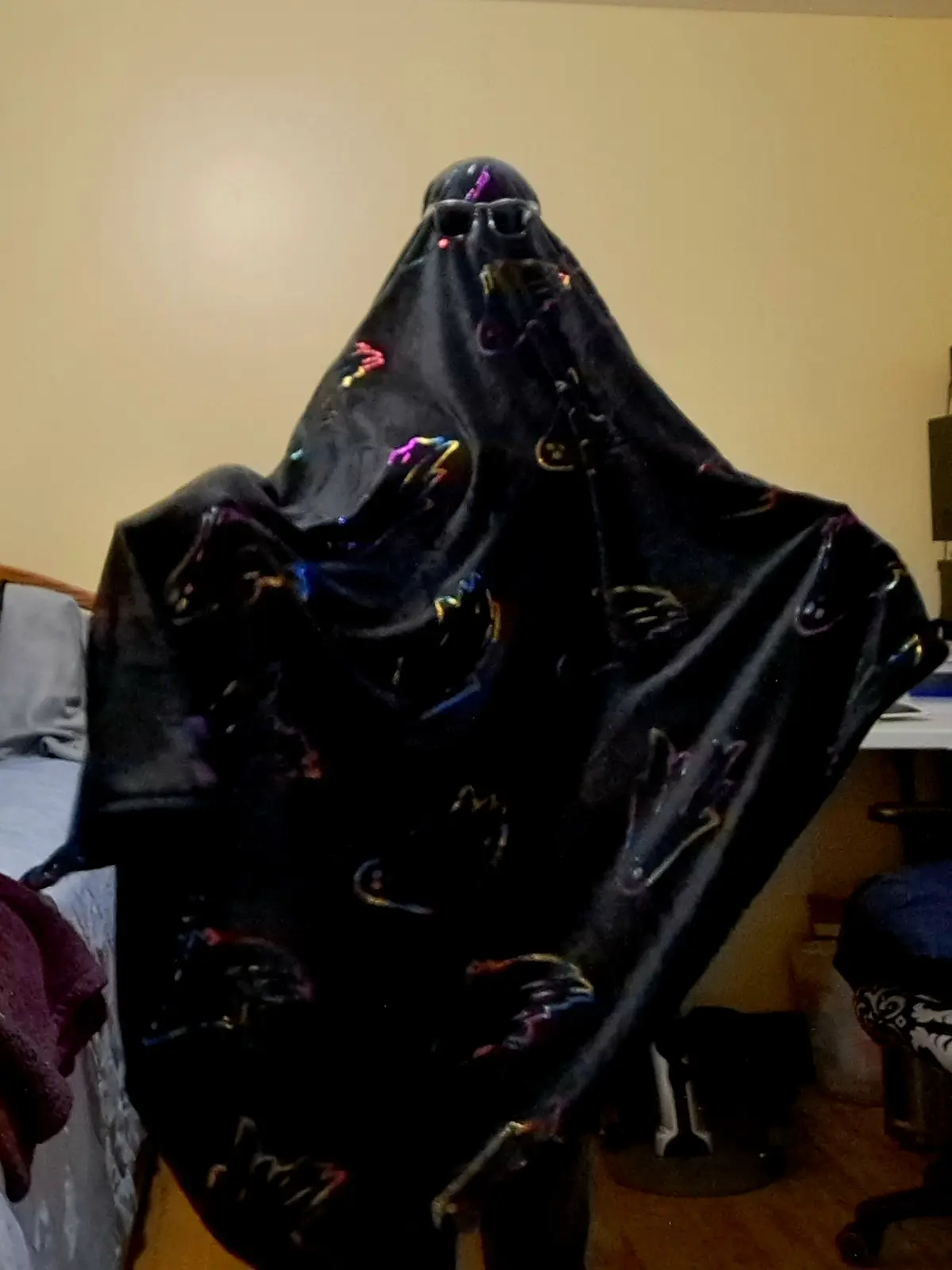A person is seated indoors, completely covered by a black blanket or cloth that drapes over their head and body, leaving…