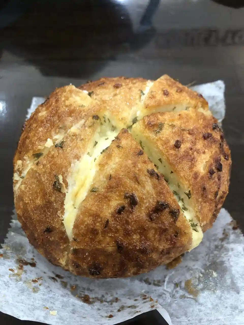 A golden-brown, round baked good with visible herbs and melted cheese oozing from its scored top sits on parchment paper…