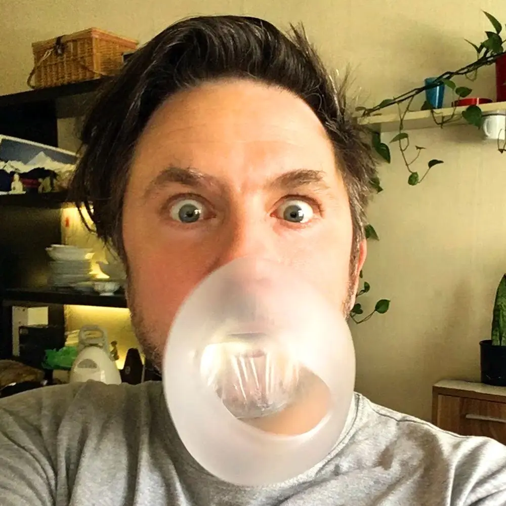 A man with wide, startled eyes is blowing a large, translucent bubblegum bubble that covers his mouth and nose. He has dark…