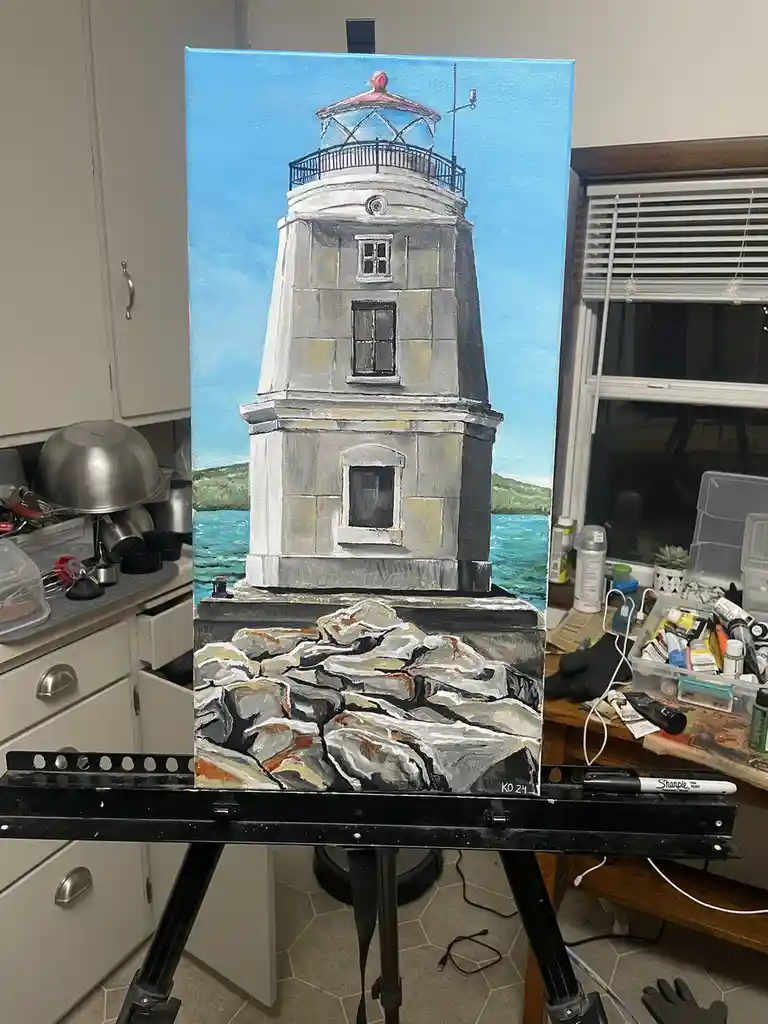 A painting of a lighthouse on a rocky shore under a blue sky is displayed on an easel in a cluttered room. The painting…