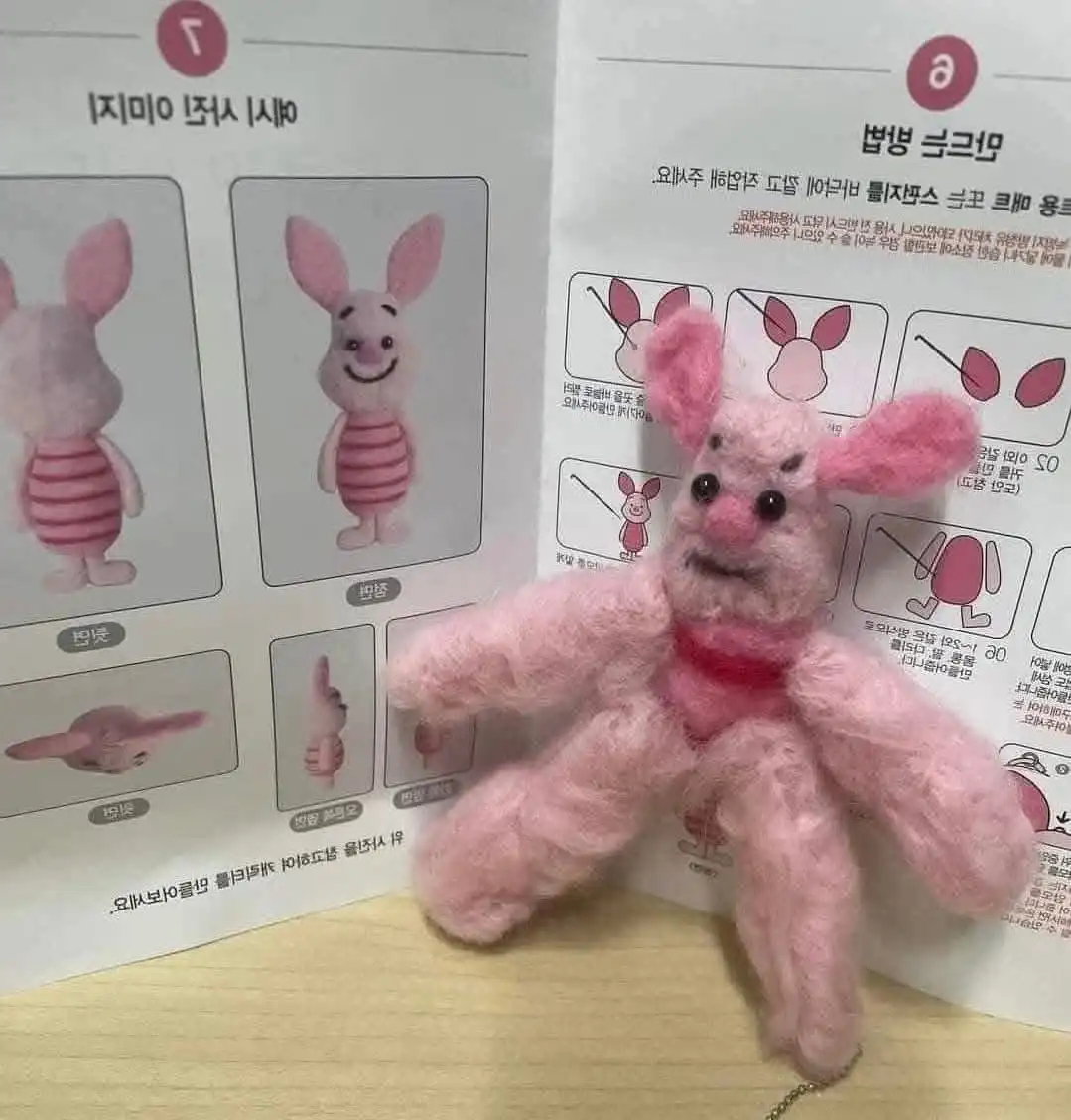 A handmade, fuzzy pink plush toy resembling a rabbit with a mischievous, slightly angry expression sits in front of a…