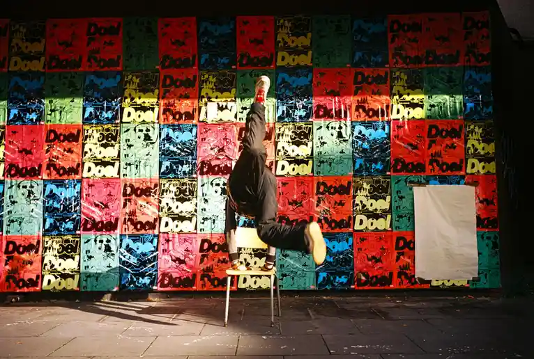 A person is upside down on a chair in front of a colorful wall covered in repeating 'Doel' posters. The image has a strong…