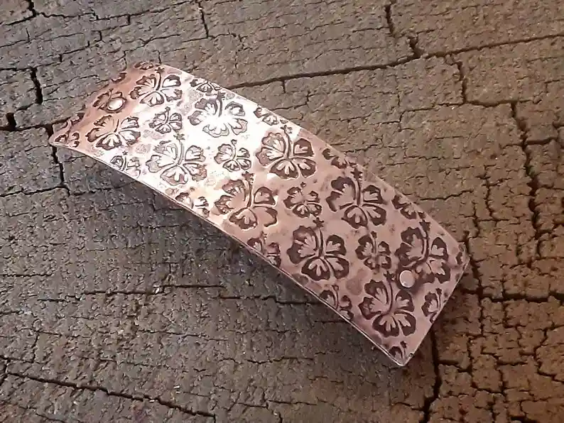 A copper-colored, rectangular bracelet with embossed hibiscus flower patterns rests on a cracked, textured surface…