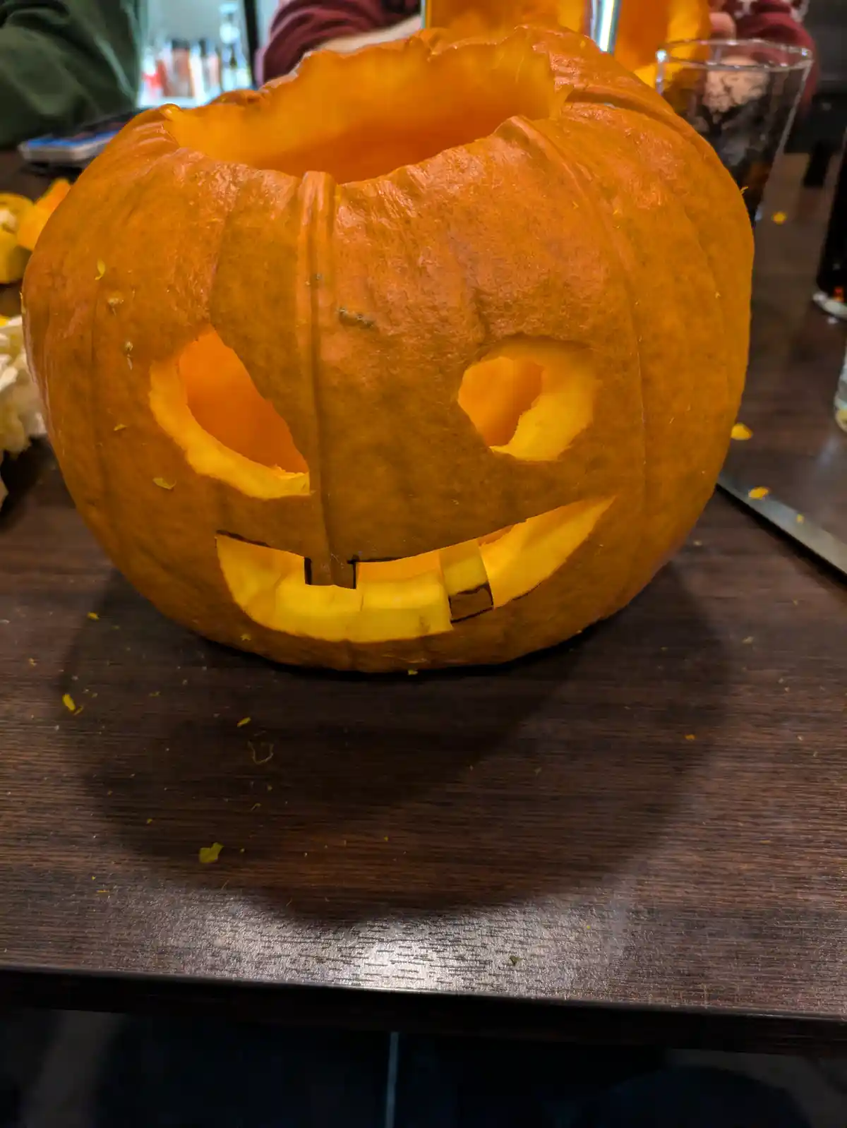 A close-up photo shows a carved orange pumpkin with a simple face, sitting on a dark wooden table. The pumpkin has two eye…