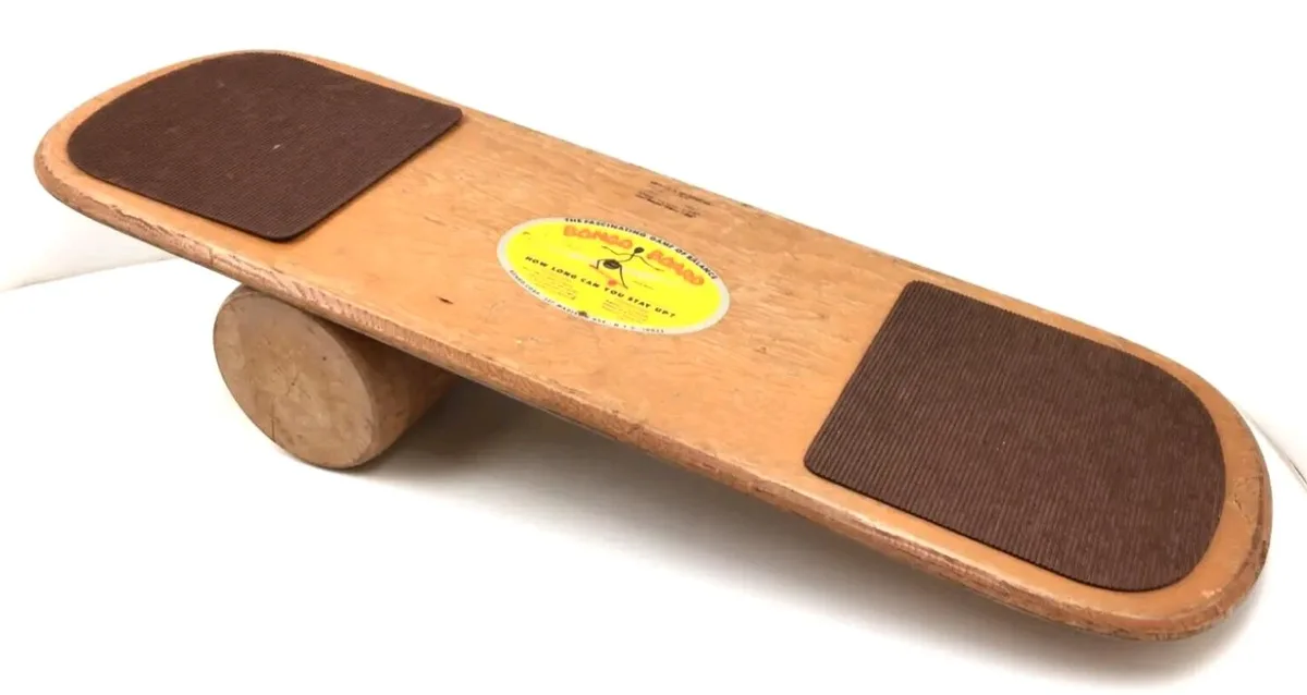 A wooden balance board toy with brown textured foot pads and a yellow label that reads 'BONGO BOARD THE FASCINATING GAME OF…