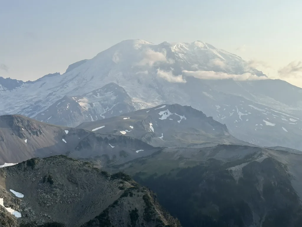 A scenic mountain landscape featuring a large snow-capped peak in the distance, partially obscured by clouds, with rugged…