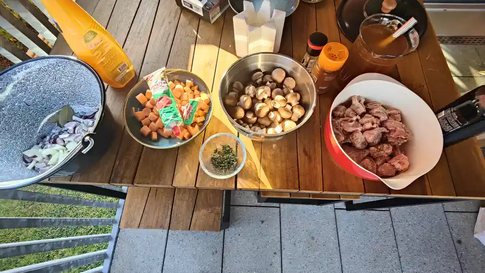 An overhead shot of a wooden outdoor table laden with ingredients for cooking: a bowl of raw meat chunks, a metal bowl of…