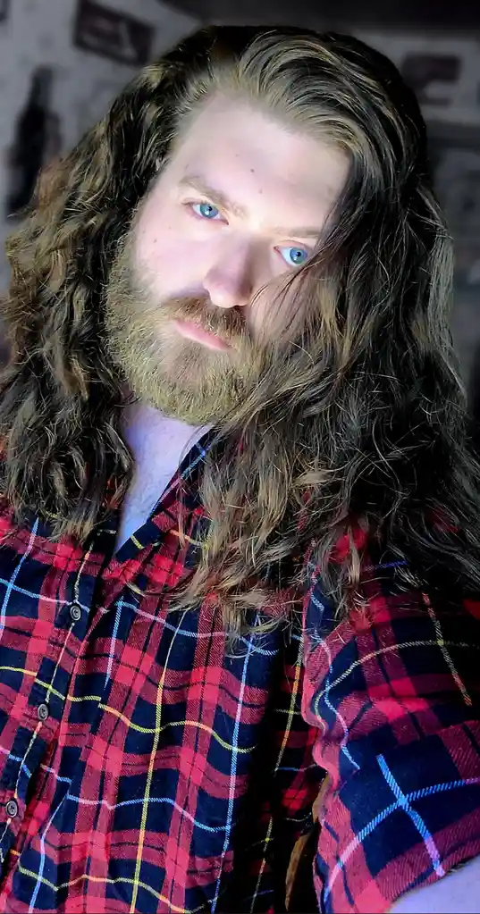 A man with long, wavy brown hair and a full beard stares directly at the camera with a serious expression, wearing a red and…