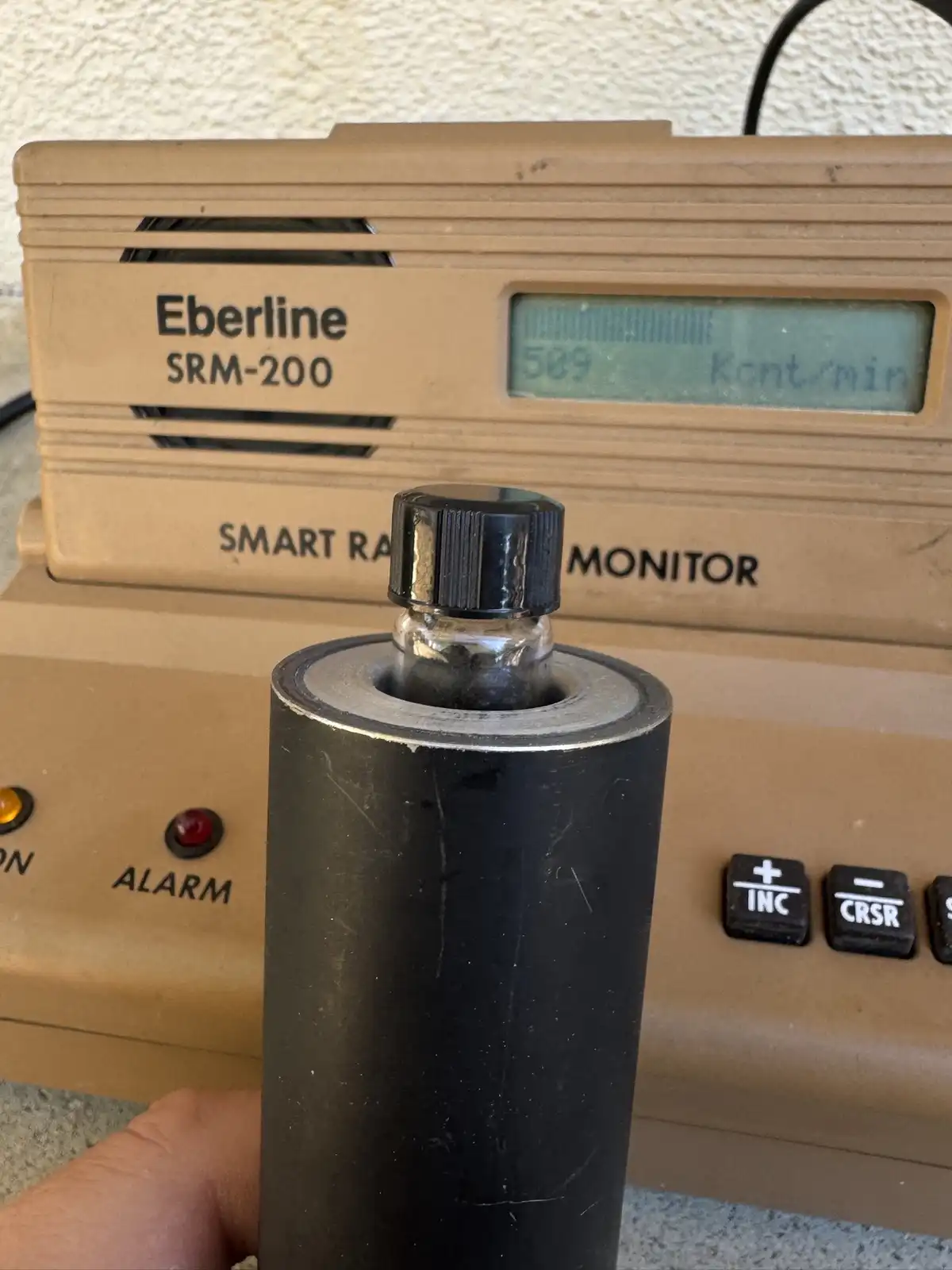 A tan Eberline SRM-200 Smart Radiation Monitor displays '589 Kont/min' on its screen, with a hand holding a black…