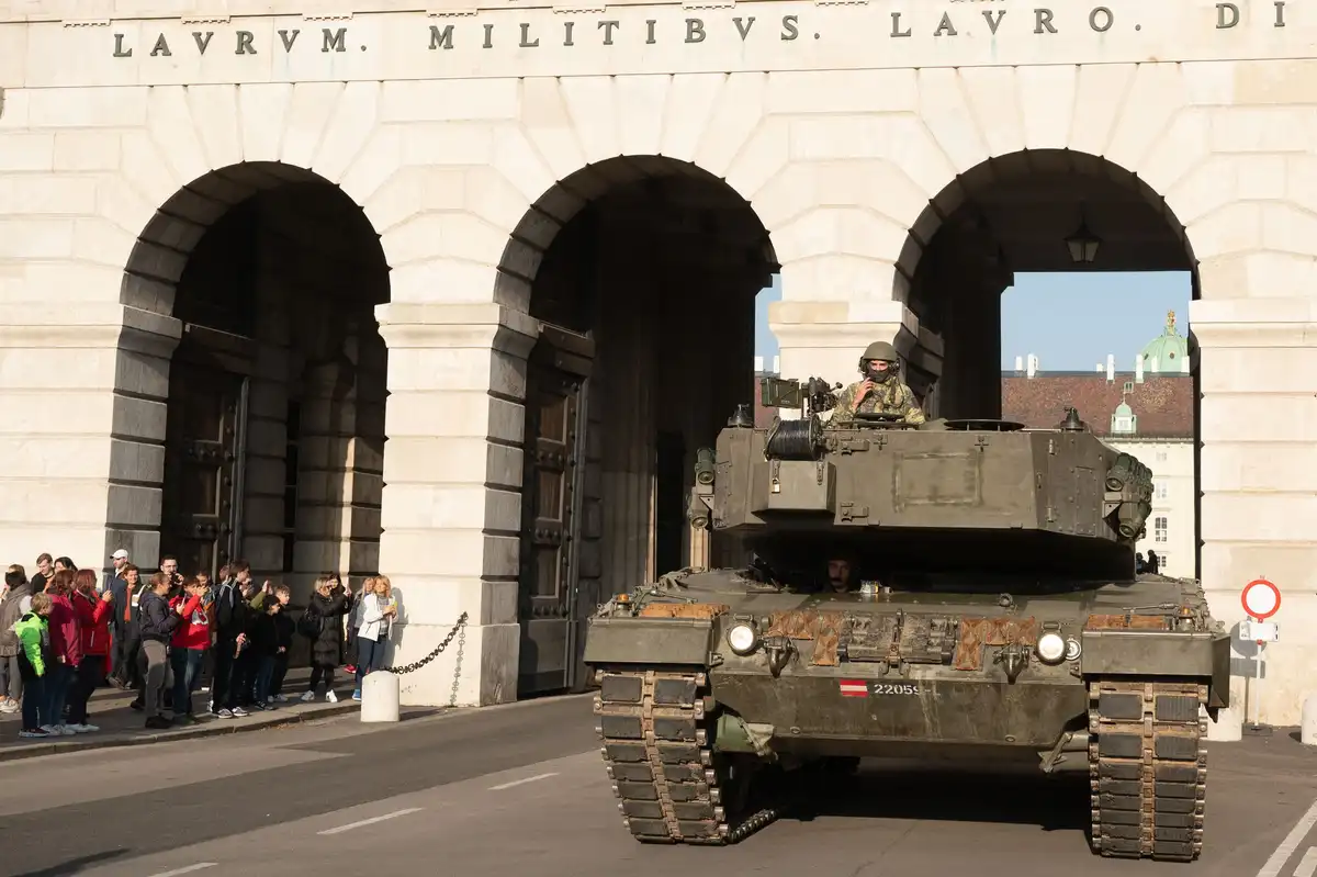 A military tank drives through a large stone archway with the Latin inscription 'LAURVM. MILITIBVS. LAURO. DI' above it…
