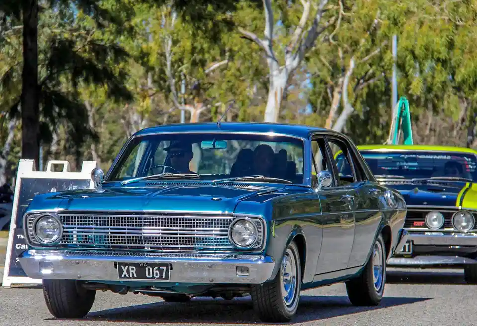A shiny blue classic car with license plate 'XR 067' is parked outdoors with trees in the background. Another vintage car is…