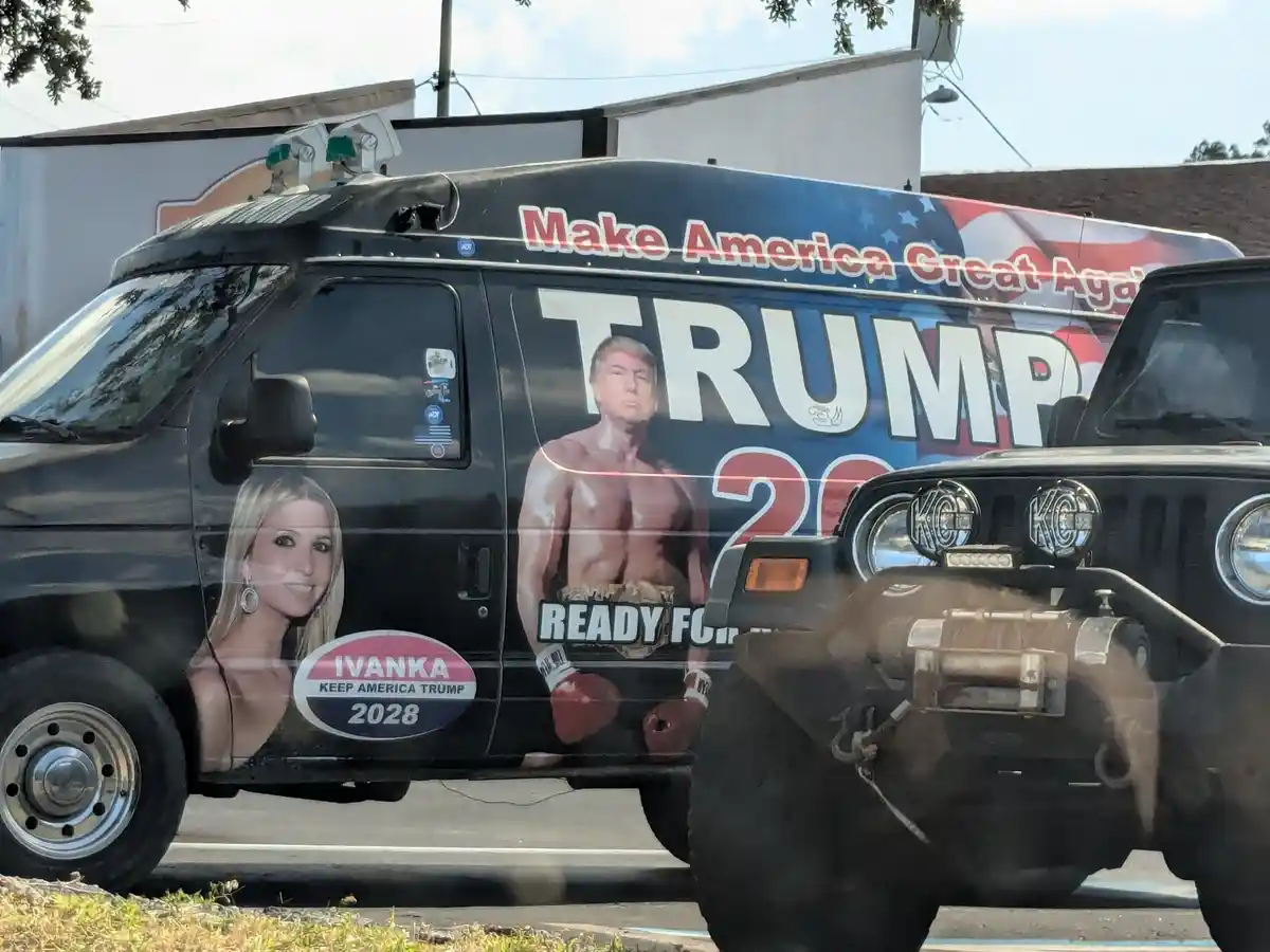 A black van is covered in political campaign graphics, featuring Donald Trump as a shirtless boxer with 'TRUMP 20' and…