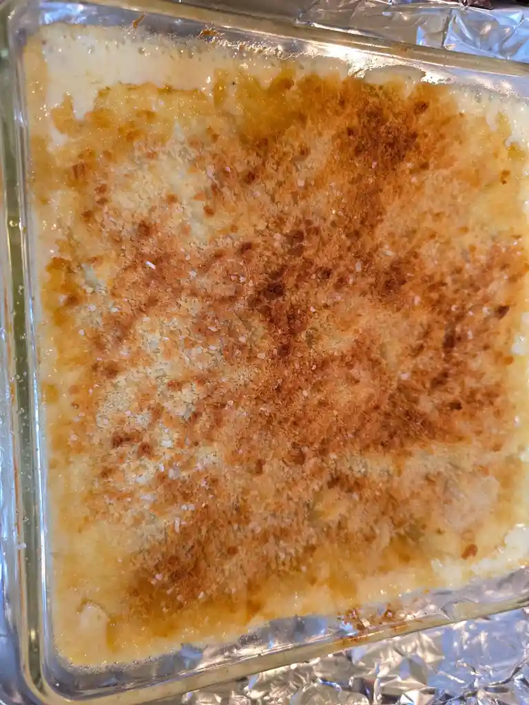 A close-up photo shows a rectangular glass baking dish containing a casserole with a golden-brown, crispy top layer…