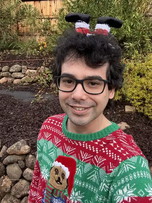 A man with curly black hair and glasses smiles while wearing a red and green Christmas sweater with a pug in a Santa hat on…