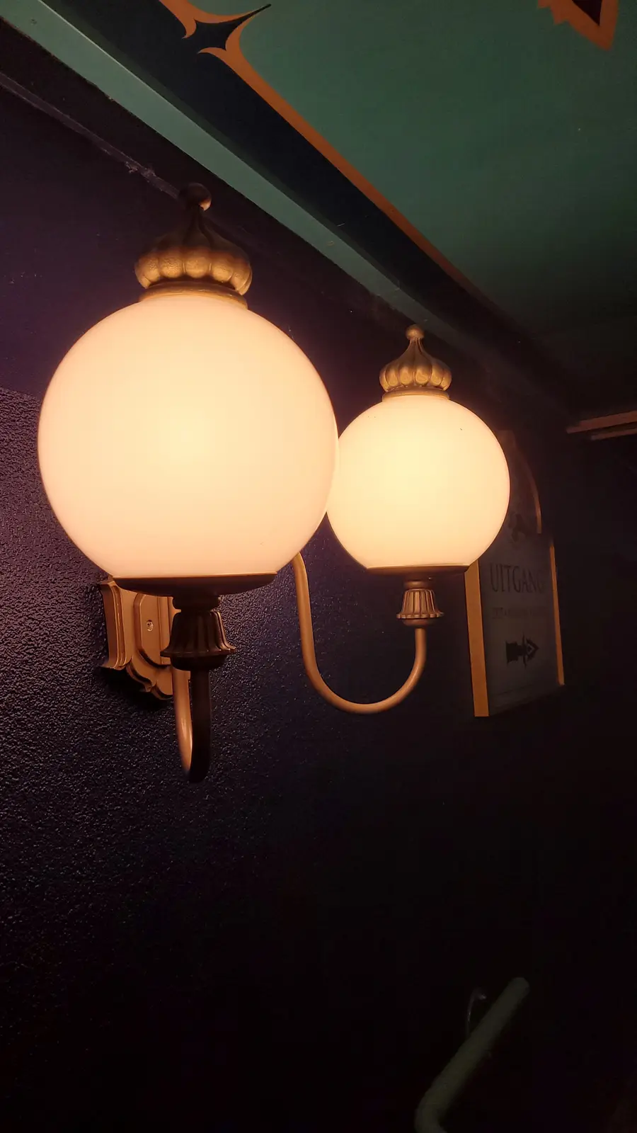 Two glowing, spherical wall-mounted lamps with brass fixtures are mounted side-by-side on a dark textured wall, next to a…