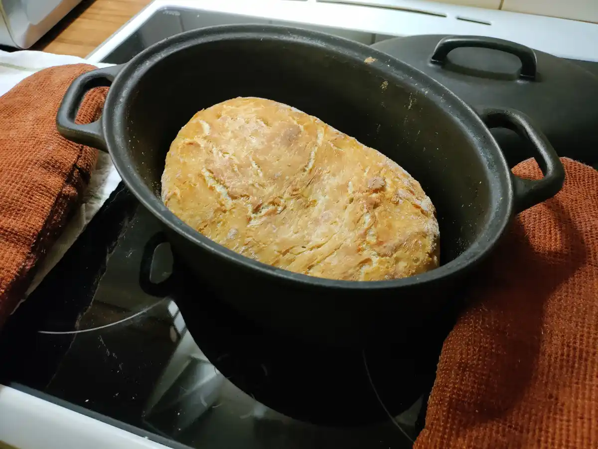 A golden-brown loaf of bread sits inside a black cast-iron Dutch oven, which rests on a black glass stovetop. Orange kitchen…