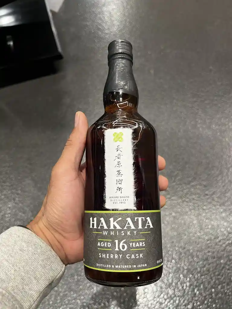 A hand holds a bottle of Hakata Whisky, aged 16 years in a sherry cask, against a textured gray background. The label…