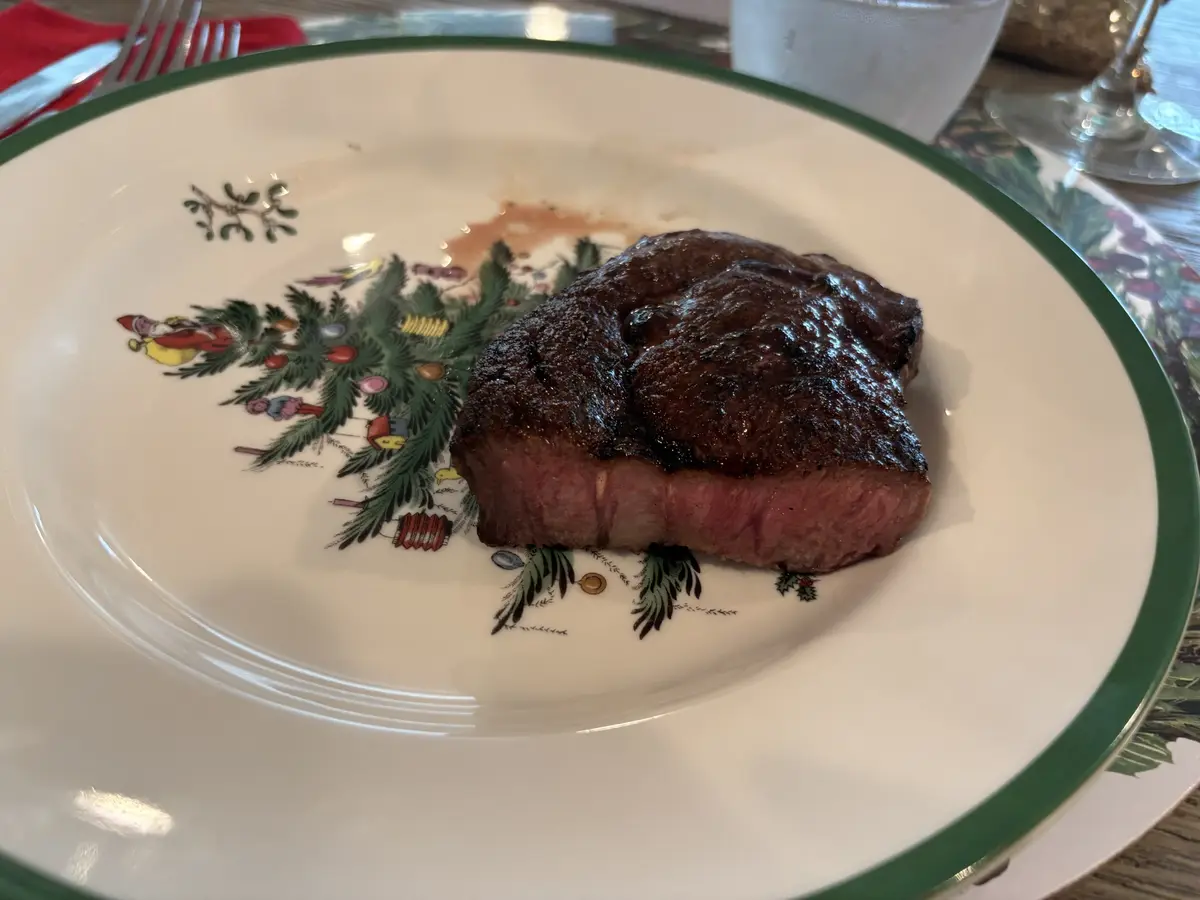 A close-up photo shows a seared, medium-rare steak resting on a white ceramic plate with a green rim. The plate is decorated…