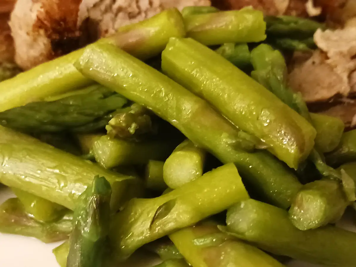 A close-up photo of cooked asparagus spears on a plate, with some pieces showing a glossy sheen from oil or butter, and…