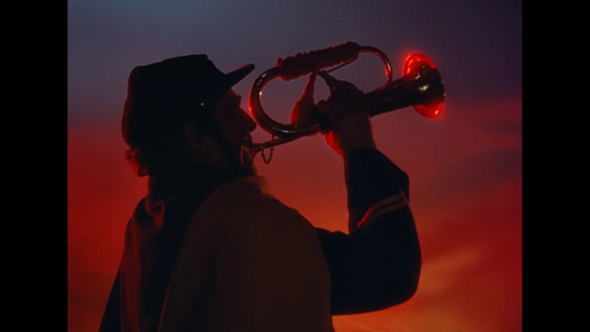 A person in silhouette, wearing a cap and jacket, plays a brass bugle against a dramatic red and blue sky. The bugle's bell…