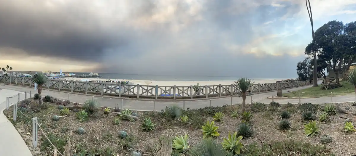 A wide-angle photo of a beach scene with a large, ominous dark cloud looming over the ocean and pier in the distance, while…