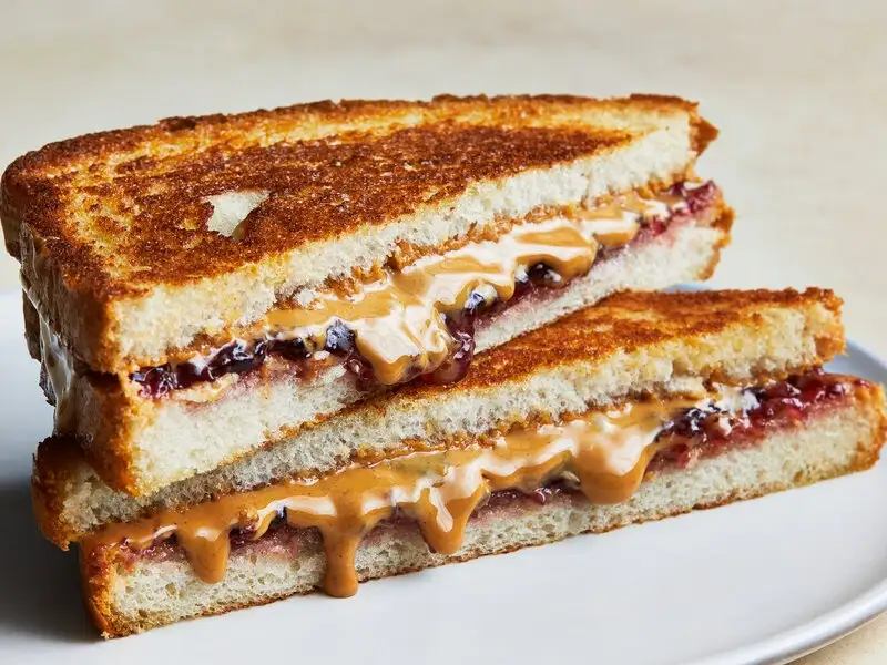 A close-up of a grilled peanut butter and jelly sandwich cut in half and stacked, with the fillings oozing out. The text…
