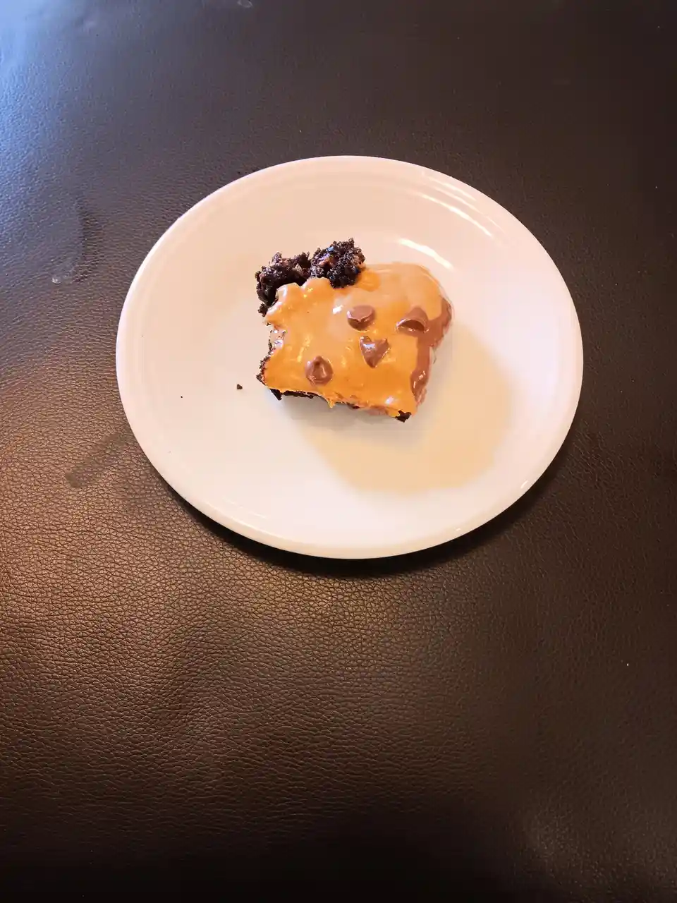 A single square piece of chocolate cake with peanut butter frosting and chocolate chips sits on a white plate, which rests…