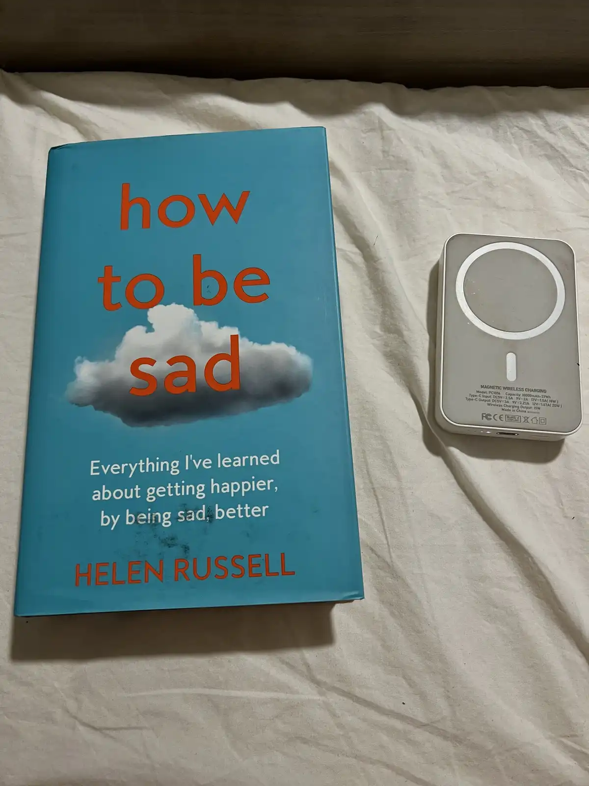 A book titled 'how to be sad' by Helen Russell lies next to a magnetic wireless charging pad on a white bedsheet. The book…