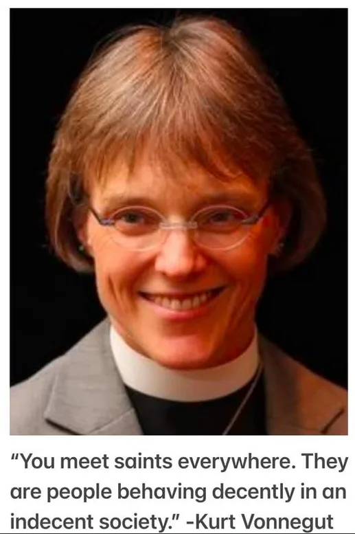 A portrait of a smiling woman with short brown hair and glasses, wearing a grey suit jacket over a black shirt with a white…