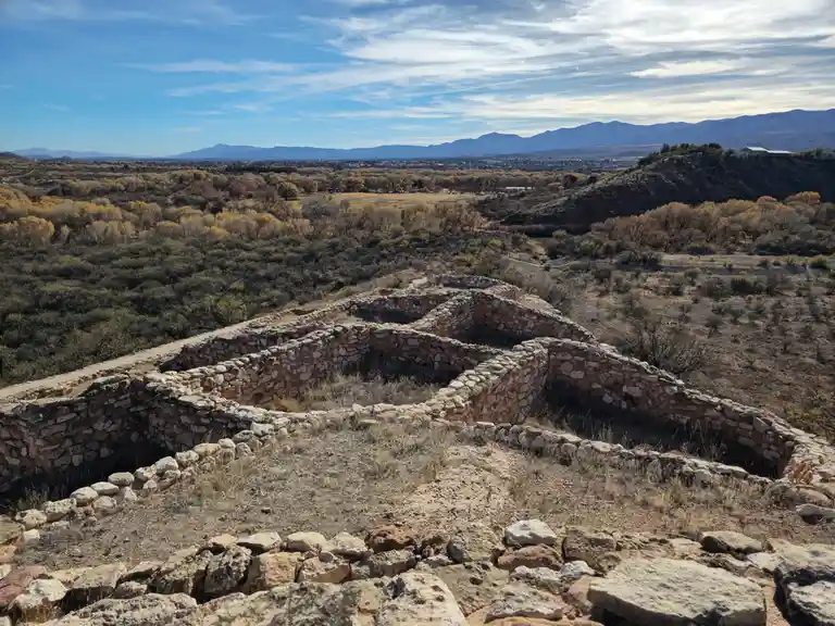 A wide-angle photo shows ancient stone ruins with low walls and courtyards, set in a dry, hilly landscape with…