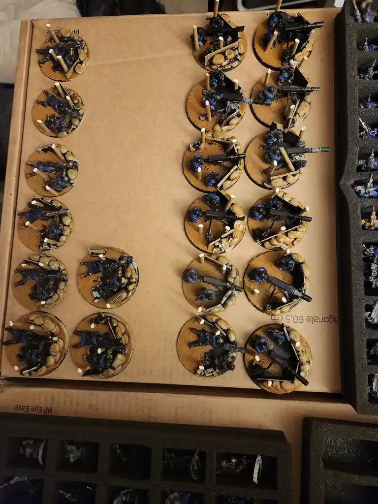 An overhead shot shows dozens of painted miniature figurines, mostly in blue and black, mounted on circular bases with…