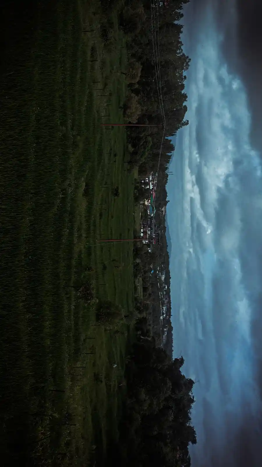 A vertically oriented landscape photo shows a dark, grassy field on the left, a distant village with lit windows on the…