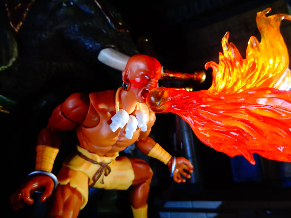 A close-up photo of a muscular action figure with a bald head, red face paint, and skull necklace, roaring while exhaling a…