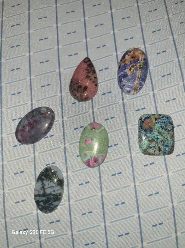 Six polished, colorful gemstones with varied patterns and holes for stringing are arranged on a blue and white striped…