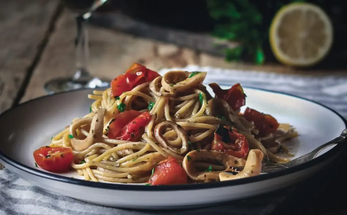 A close-up photo of a plate of pasta with mushrooms and cherry tomatoes, garnished with herbs, set on a wooden table with a…