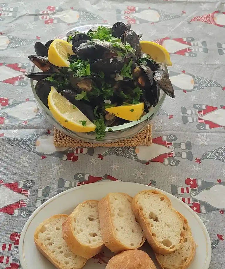 A glass bowl filled with cooked mussels, garnished with fresh parsley and lemon wedges, sits on a woven coaster atop a…