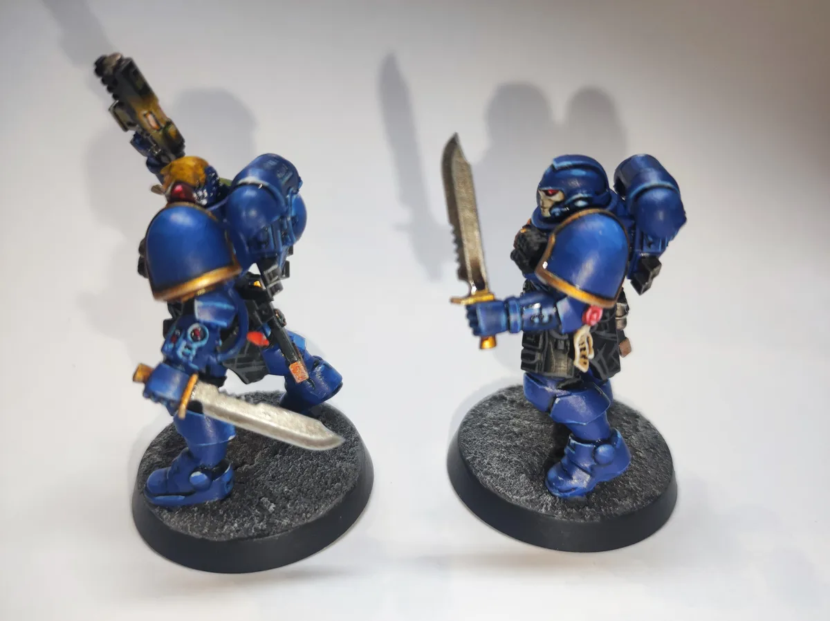 Two blue-painted miniature space marines in power armor stand on circular bases against a white background. The figure on…