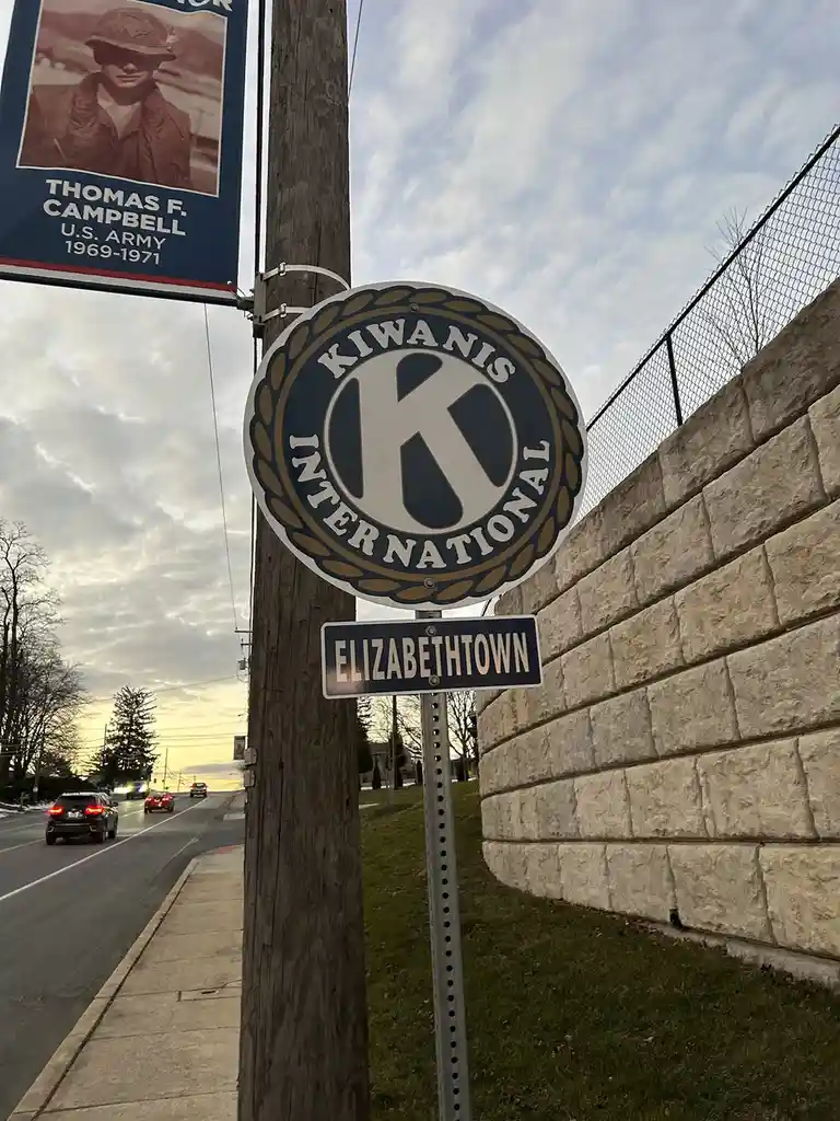 A street sign for 'ELIZABETHTOWN' is mounted below a circular 'KIWANIS INTERNATIONAL' logo sign on a wooden utility pole…