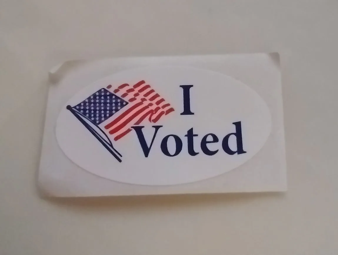 A close-up photo of a white oval sticker with a small American flag graphic on the left and the text 'I Voted' in dark blue…