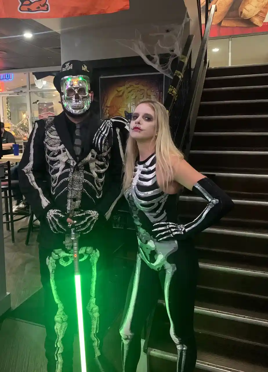 Two people dressed in full skeleton costumes pose indoors near stairs; the man wears a glowing skull mask and holds a green…