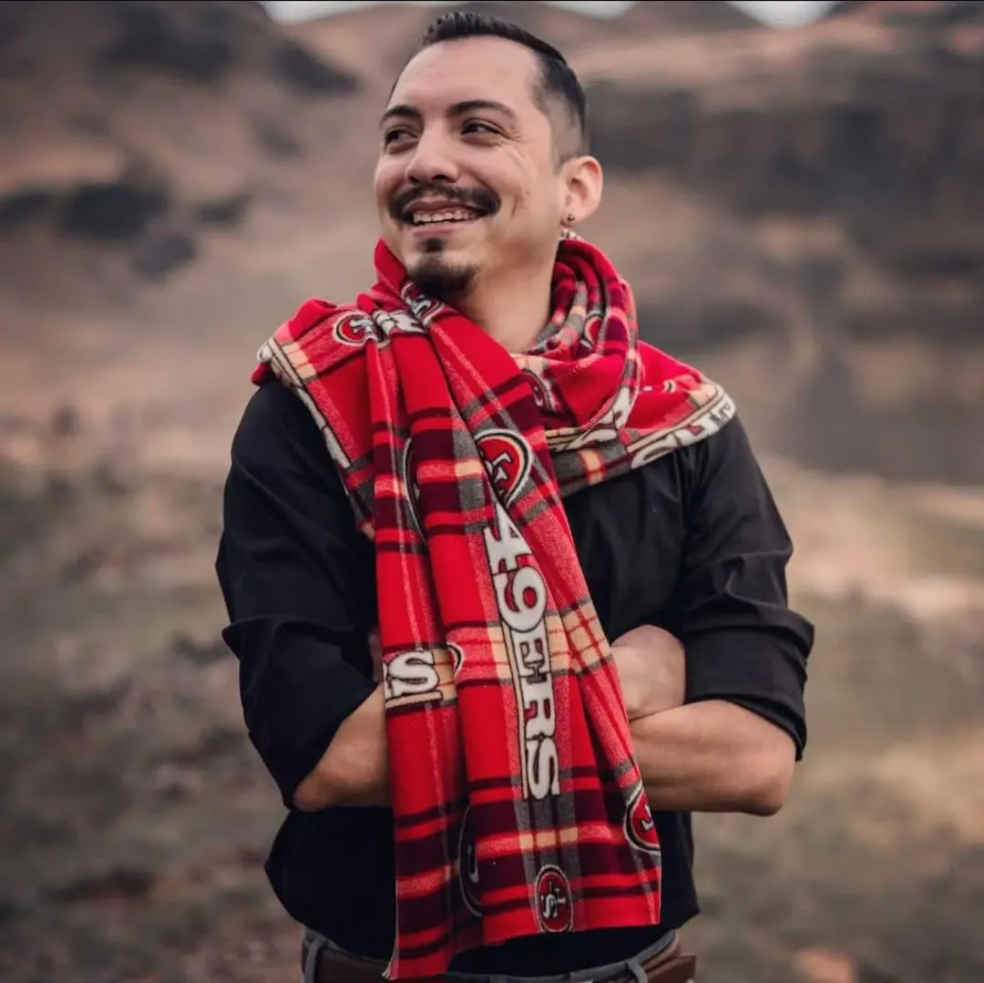 A man with a mustache and goatee stands outdoors with arms crossed, wearing a red plaid scarf featuring the San Francisco…