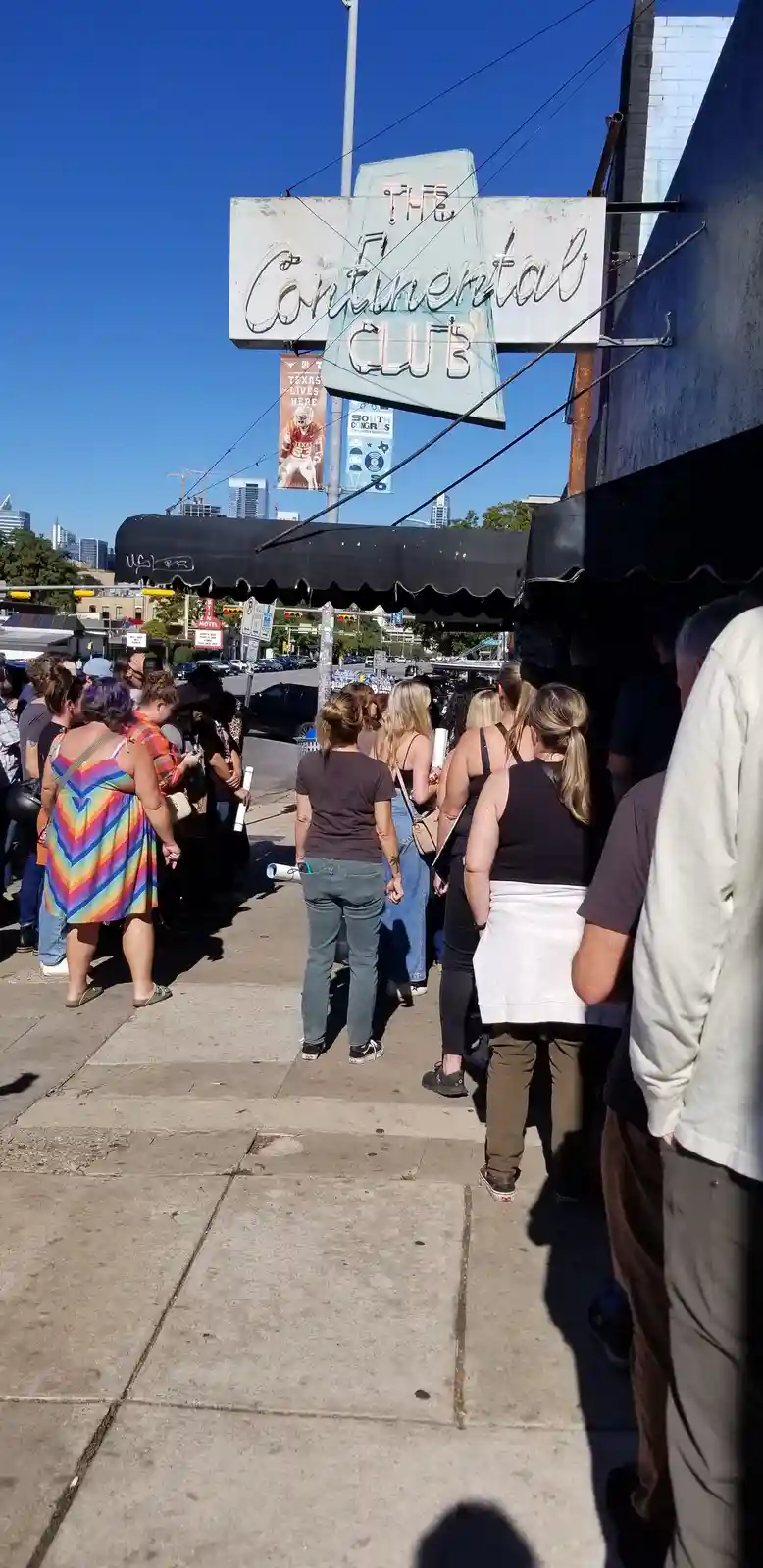 A crowd of people stands in a long line outside The Continental Club, a venue with a large, weathered sign above the…