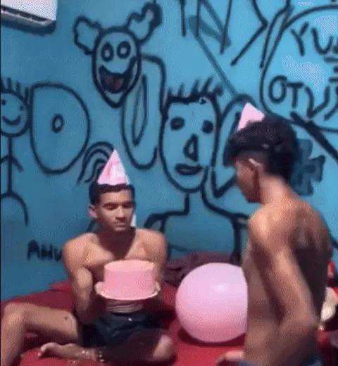 A shirtless man wearing a pink party hat sits at a red table with a small pink cake and a pink balloon. He blows out the…