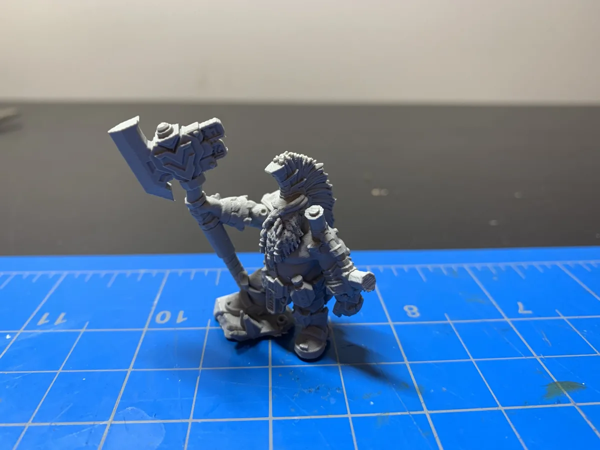 A gray, unpainted miniature figurine of a fantasy warrior with a large hammer stands on a blue grid cutting mat. The…
