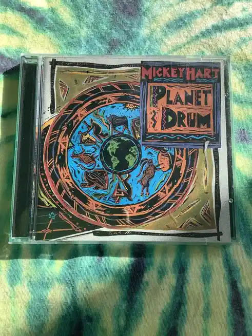 A CD case for Mickey Hart's album 'Planet Drum' rests on a tie-dye fabric. The cover art features a stylized circular design…